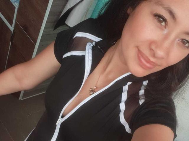 TatianaRios: Uma mulher de cabelos escuros, olhos castanhos e um sorriso, vestindo uma blusa preta com zíper e detalhes brancos, usando um colar com um pequeno coração e com um busto discreto..