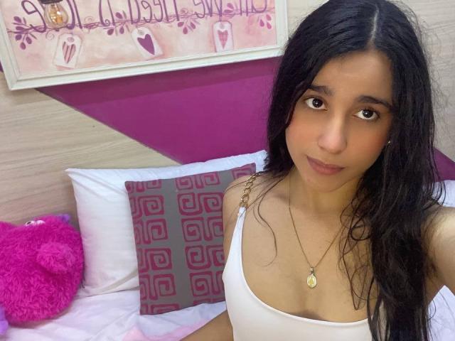 TiffannyRose, avec de longs cheveux noirs, des yeux marron et un teint hâlé, portant un débardeur blanc, avec de petites seins, et un collier en or, assise sur un lit avec des coussins roses et gris..
