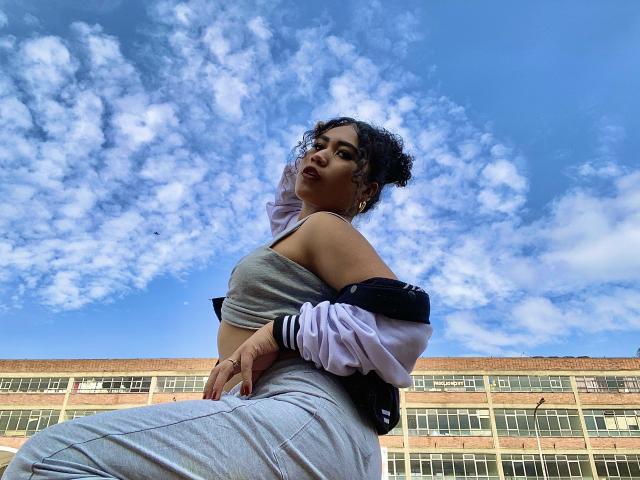 SophieSonyas with curly hair, wearing a grey sports bra and light grey pants, has a black and white jacket off her shoulders, looking up at a blue, cloudy sky.