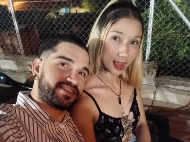 SophiaAndDaimonX with blonde hair, small tits, black lace top, tongue out, next to DaimonX, bearded, striped shirt, earring, chain necklace, both smiling, outside at night.