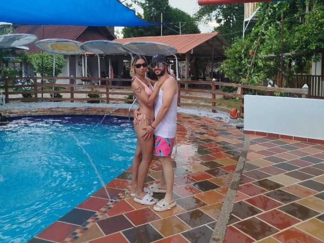 SophiaAndDaimonX stand by a pool, she's topless with small tits, wearing a thong, he's in pink and blue swim trunks, both with sunglasses, arms around each other.