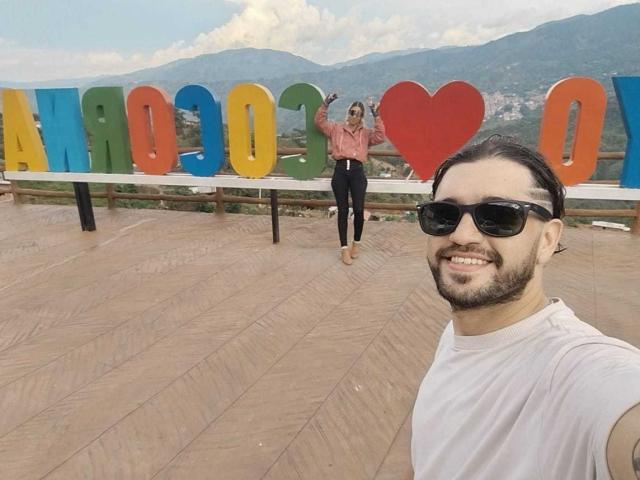SophiaAndDaimonX, wearing sunglasses, white tee, and black pants, smiling with beard, long dark hair, and white skin, stands in foreground; SophiaAndDaimonX, waving, in background with colorful heart.