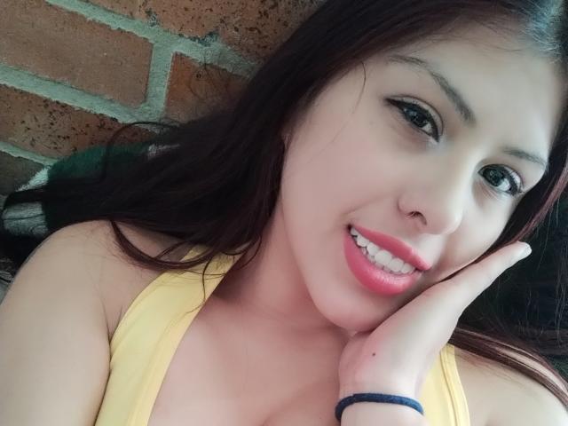 ElaiaaBakerr has pink lips, white teeth, and dark brown hair, smiling with a hand on her cheek. She's wearing a yellow top, with a black bracelet on her wrist.
