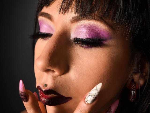 TerriClement's face with purple eyeshadow, dark lipstick, green nose piercing, white and marble nail, pink nail, black hair, and hoop earrings, eyes closed, dark background.