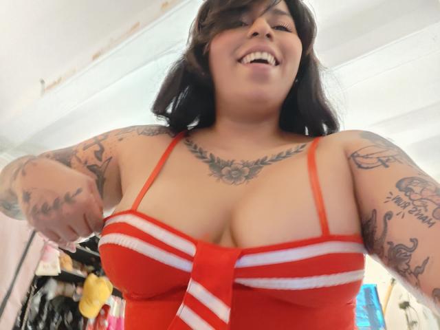 Chili, with medium-sized breasts and tattoos, wears a red and white striped top, smiling while adjusting her bra straps.