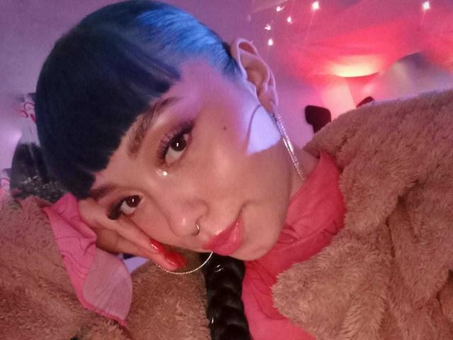 GigiVolkova, con cabello corto azul, rostro brillante, aro en la nariz, aretes de aro, camisa rosa, chaqueta marrón esponjosa, uñas rojas, acostada, iluminación rosa y púrpura, mirando a la cámara..