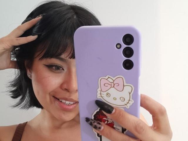 JesicaRossi with short black hair, smiling, holding a purple phone with a Hello Kitty sticker, wearing a brown top, and showing off black nails.