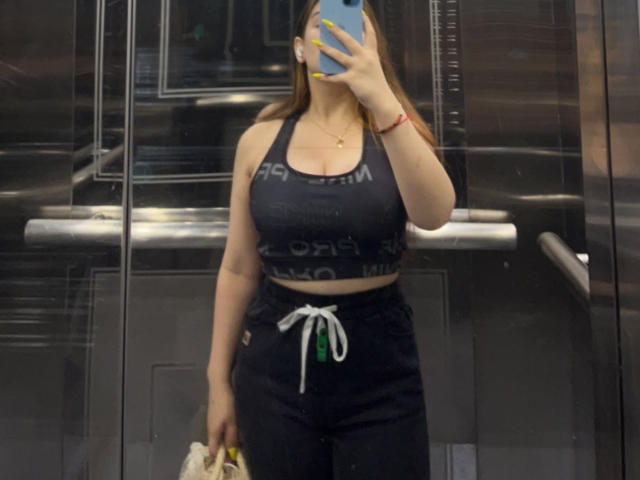 BeverlyApple, con grandes senos, vistiendo un top deportivo negro y pantalones negros con un cordón blanco, sosteniendo un bolso y un teléfono, selfie frente al espejo en un ascensor metálico..