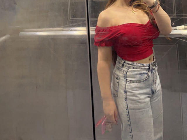 BeverlyApple con una blusa roja de corte asimétrico que deja ver sus pechos, y jeans azules de cintura alta, sosteniendo un cepillo de pelo rosa, en un selfie frente al espejo..