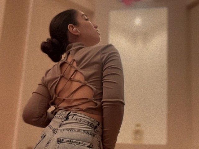 BeverlyApple staat met haar rug naar de camera, ze draagt een bruin top met lange mouwen en een kruisende rug, en lichtblauwe jeans met een hoge taille. Haar donkere haar zit in een knot..