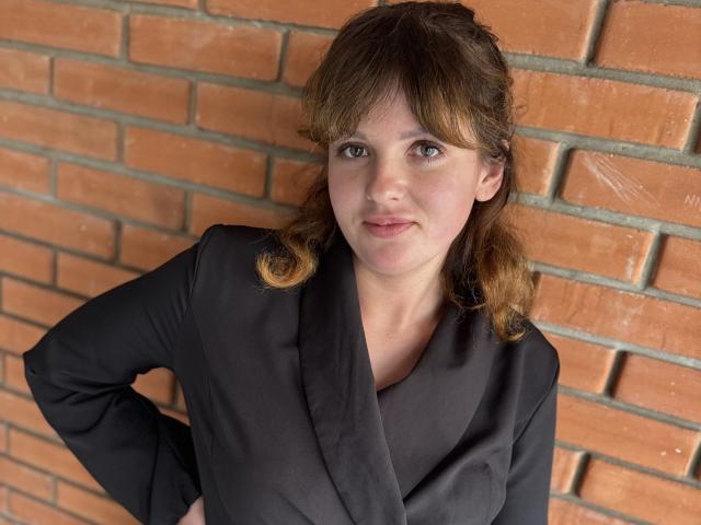 StaceyLex has light brown hair, wearing a black button-up shirt with a cowl neck, hands on her hips, looking at the camera with a slight smile.