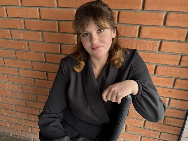 StaceyLex with wavy brown hair, wearing a black button-up shirt, sitting in a chair, her right arm resting on the chair's armrest, smiling slightly.