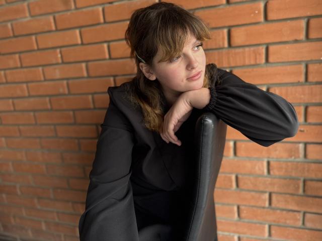 StaceyLex met bruin haar, gekleed in een zwarte jurk, leunt tegen een stoel, armen gekruist, kijkt opzij, een bakstenen muur op de achtergrond, zachte uitdrukking, lichte glimlach..