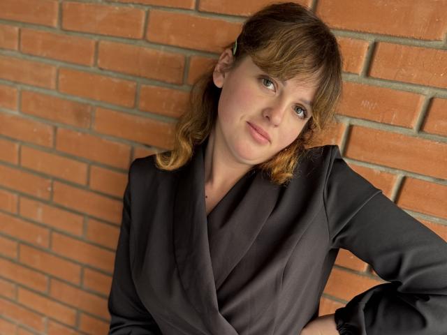StaceyLex, met golvend bruin haar, draagt een zwarte satijnen blouse en leunt tegen een bakstenen muur. Ze kijkt opzij met een lichte glimlach, waarbij haar zachte, lichte huid zichtbaar is..