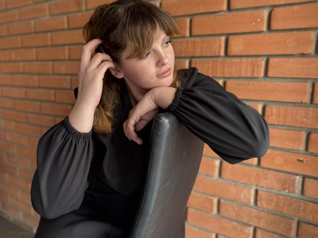 StaceyLex, com cabelo castanho, vestindo um vestido preto com mangas bufantes, está sentada em uma cadeira preta, com a mão no cabelo, olhando para o lado, com uma parede de tijolos ao fundo..