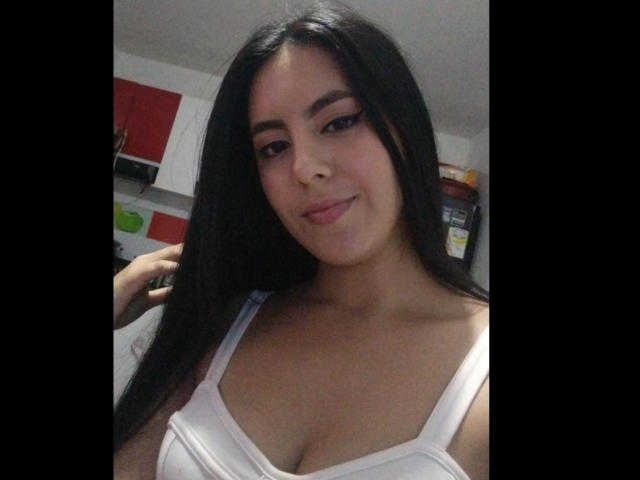 EimmyPark, com cabelos longos e pretos, vestindo uma camiseta branca, mostrando seus seios, sorrindo levemente, em um quarto com pouca luz. Sua maquiagem inclui delineador escuro e batom rosa..