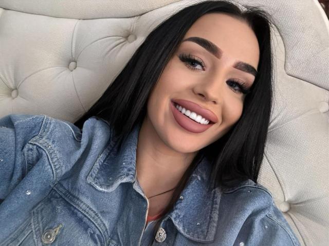 EvaHotRose mit langen schwarzen Haaren, vollen rosafarbenen Lippen und weißen Zähnen, trägt eine blaue Jeansjacke und lächelt dabei leicht mit der Zunge heraus. Sie liegt auf einem weißen, gepolsterten Kissen..