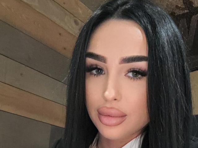 EvaHotRose hat schwarzes Haar, volle Augenbrauen, lange Wimpern und rosafarbenen Lippenstift. Sie schaut direkt in die Kamera und lächelt leicht und selbstbewusst. Ihre Haut ist hell..