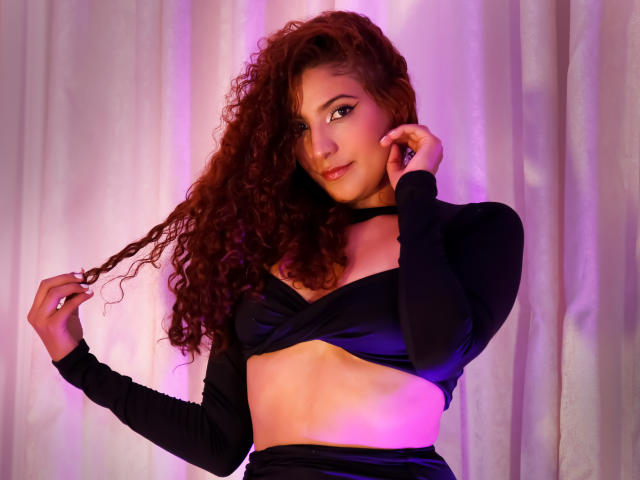 MiaBianchi with curly red hair, wearing a black crop top and long sleeves, holding her hair, showing her small tits and toned midriff, with a slight purple glow.