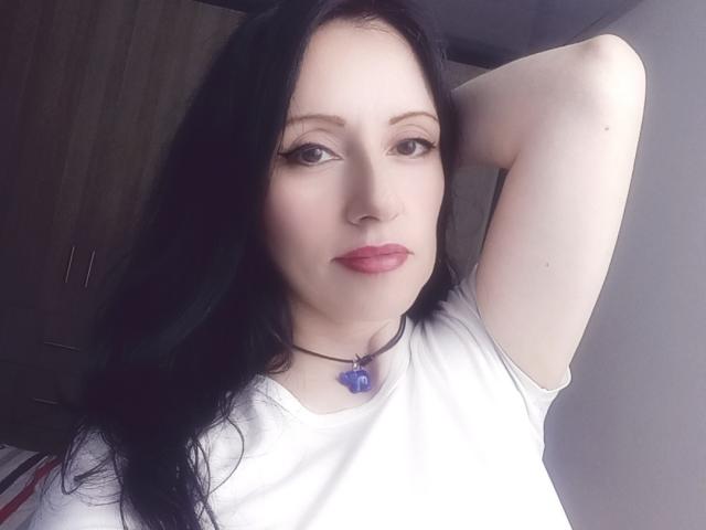 MondBlue with pale skin, black hair, and pink lips, wearing a white shirt, has her arm raised, showing her armpit, and a purple heart necklace.