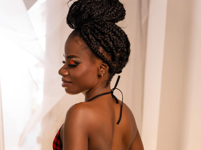 MiaHopson with dark skin, braided black hair in a bun, orange eyeshadow, black halter top, showing her back and ass, looking to the side, soft light, calm expression.