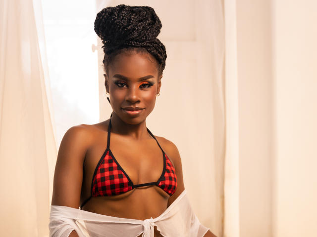 MiaHopson with dark skin and braided hair wearing a red and black checkered bikini top, white sheer shirt off her shoulders, smiling subtly, standing in soft light.
