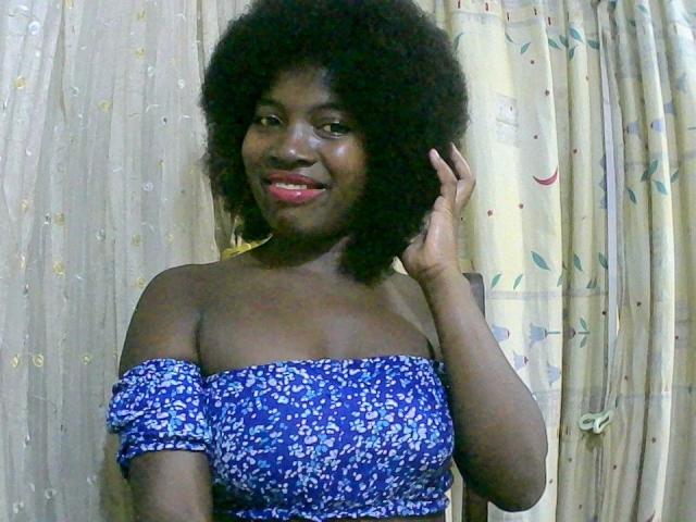  AlexandraC with dark skin, curly black hair, red lipstick, wearing a blue floral off-shoulder dress, smiling, hand touching her hair, showing her bare shoulders and small tits.