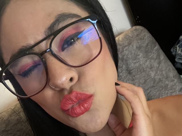 StellaBeckker with pink eyeshadow, black eyebrows, thick glasses, red lipstick, black hair, and a nose ring, puckering her lips, hand near her neck.