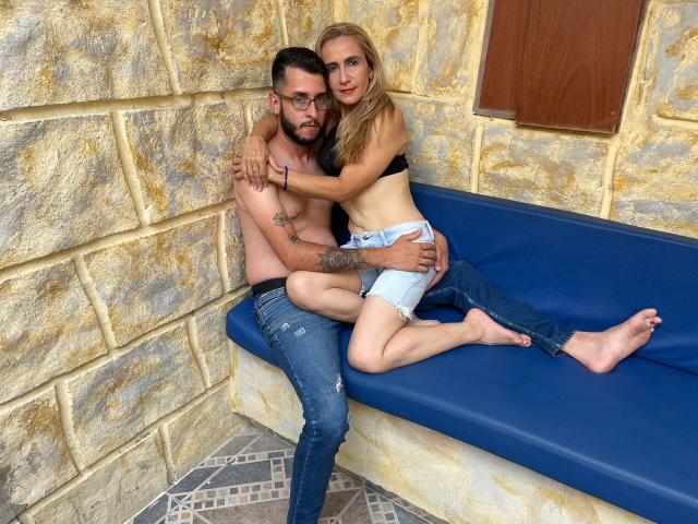 TifanyAndStiven sitting on a blue bench, TifanyAndStiven with tits in a black bra, jeans down, Stiven shirtless, tattooed, holding her ass, both looking at camera.