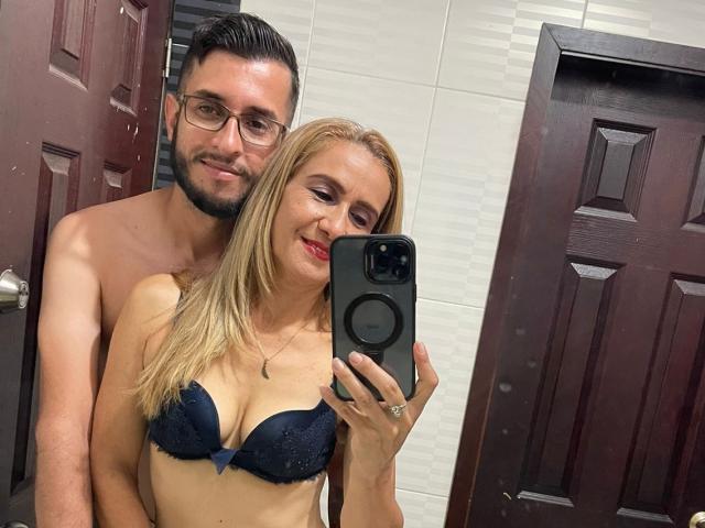 TifanyAndStiven standing in a bathroom, Tifany with blonde hair, red lipstick, and navy lace bra, Stiven with glasses, beard, and brown hair, both smiling at the mirror.