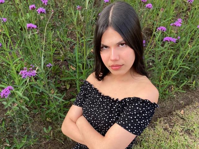 AmyXHottie with dark hair, crossed arms, and a serious face, wearing a black off-shoulder polka dot dress, standing in front of purple flowers and green grass.