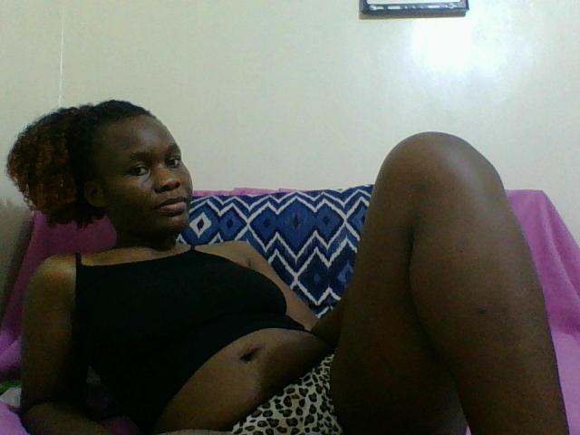 DeelaHottie with dark skin, curly red-tinted hair, black tank top, and leopard print shorts, reclining on a pink-covered couch, leg raised, looking at the camera.
