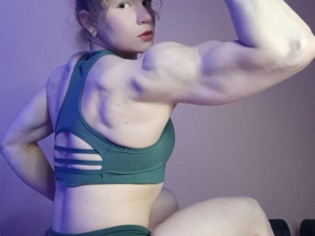 VeraKyk with muscular arms, flexing, wearing a green sports bra with cutouts, showing her back and butt, in a room with purple lighting. Her face is slightly turned.