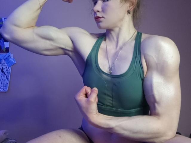 VeraKyk flexing her muscular arms, wearing a green sports bra, showing off her bulging biceps and strong, defined muscles. She has pale skin and a serious expression.