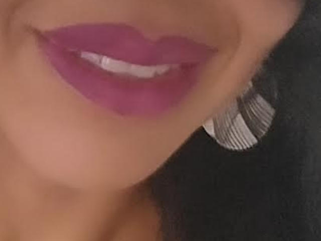 FlorLuna69s glänzende, violette Lippen und ein Hauch von weißen Zähnen, mit einem dunklen Ohrring und schwarzen Haaren, in einem leicht unscharfen Selfie..