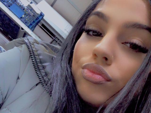 CherryDevine's face with long black hair, full pink lips, and thick black eyelashes, wearing a puffy grey jacket, in a selfie. Her nose is pierced on the right side.