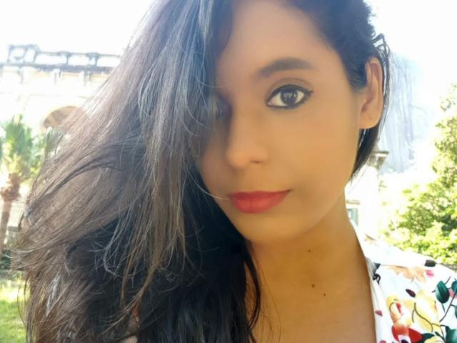 Caffein has long black hair, one eye closed, red lipstick, and a floral shirt, with a smooth, tan face and subtle makeup. She's looking at the camera.