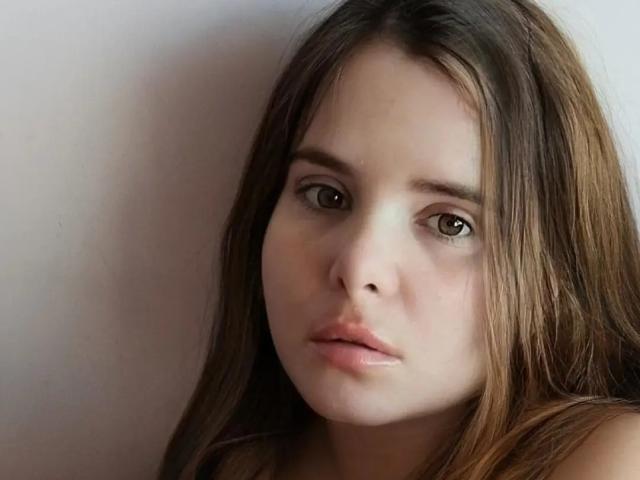 Dolly69X has long brown hair, brown eyes, and a neutral expression with slightly parted pinkish lips. She's looking directly at the camera with a soft, natural light.