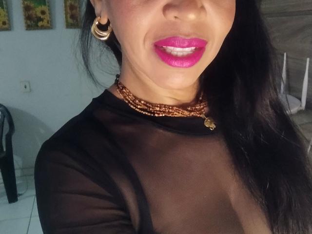 FlorLuna69 with shiny pink lipstick, black sheer top, gold necklace, and hoop earrings, showing her tits through transparent fabric. Her brown skin and long black hair are visible.