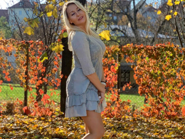 Lea Diamond, con cabello rubio, sonriendo, vestida con un vestido azul de encaje, de pie en un jardín otoñal iluminado por el sol, sosteniendo una taza blanca, con las manos sobre su pecho, rodeada de hojas amarillas..