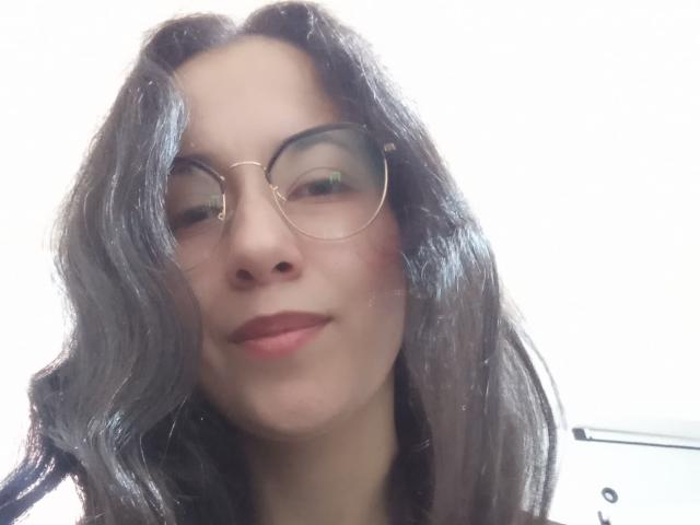 AnneStone with wavy black hair, wearing round glasses, slight smile, and pinkish-red lipstick, looking down, soft light, white background, visible part of a laptop on the right.