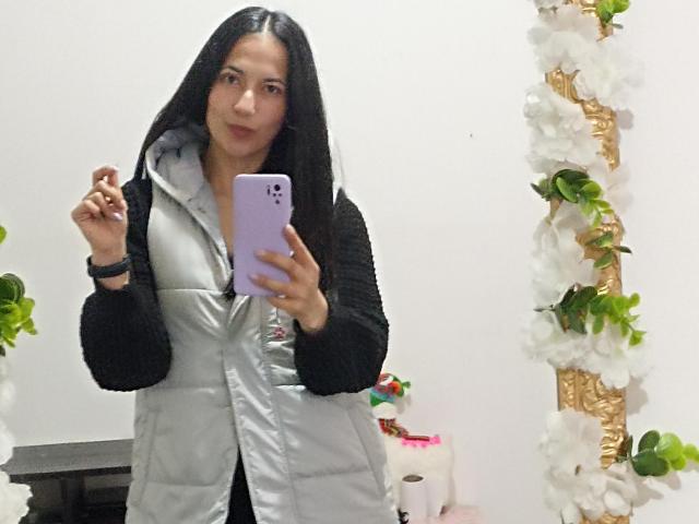 AnneStone with long black hair, wearing a grey puffy vest over a black hoodie, holding a purple phone, standing in front of a mirror with white floral decorations.