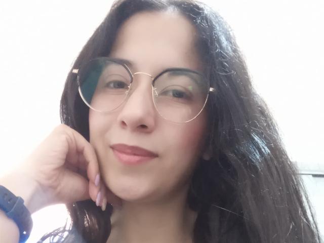 AnneStone with round glasses, dark wavy hair, pink lips, and a slight smile, resting her chin on her hand, wearing a blue wristband, looking cute.