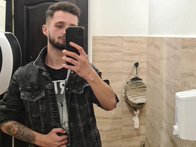 Boyka69 with short brown hair, beard, tattoo on right arm, wearing denim jacket, black shirt, taking selfie in bathroom, holding phone in right hand, toilet paper dispenser in background.