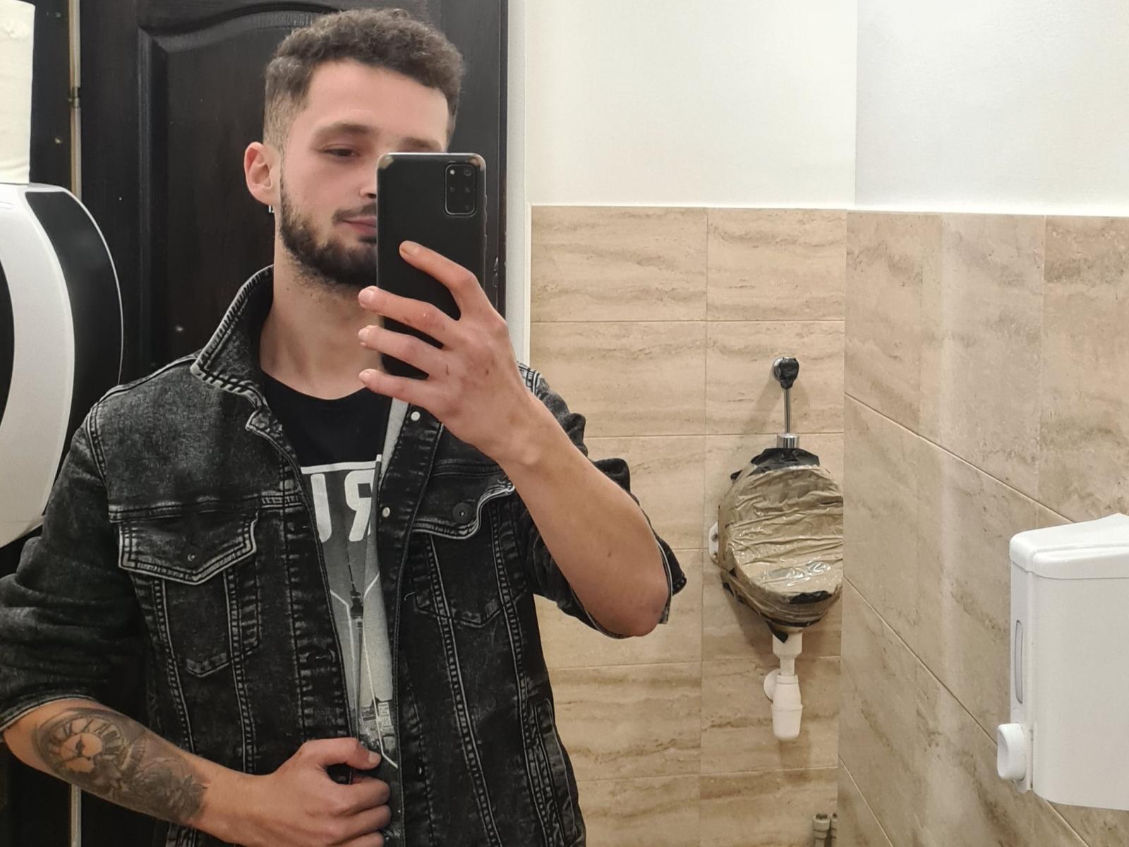 Boyka69 with short brown hair, beard, tattoo on right arm, wearing denim jacket, black shirt, taking selfie in bathroom, holding phone in right hand, toilet paper dispenser in background.