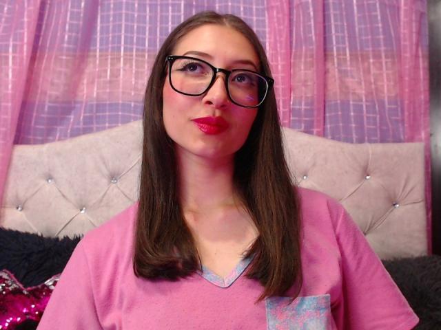 ElizabethMorrison with straight brown hair, black glasses, pink lipstick, and pink shirt, has a cute face, small tits, and a slightly chubby ass. She's standing in front of a bed.