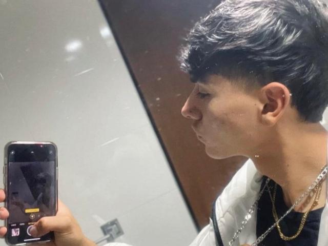 ImJuanda with short black hair, pensive expression, holding phone, wearing black shirt and gold chain, in mirror selfie. Reflection shows slight sweat on face.