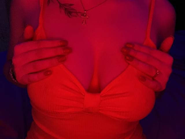 Olieri's hands on her red-dressed tits, fingers with red nails, cross necklace, tattoo on left shoulder, red lighting. Boobs, hands, nails, cross, tattoo.