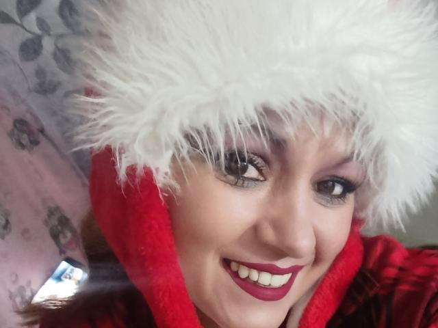 NatashaChaus with white fluffy hat, red coat, and red lipstick, smiling with visible teeth, has dark eye makeup and mascara, and is wearing a red hood. Boobs not visible.