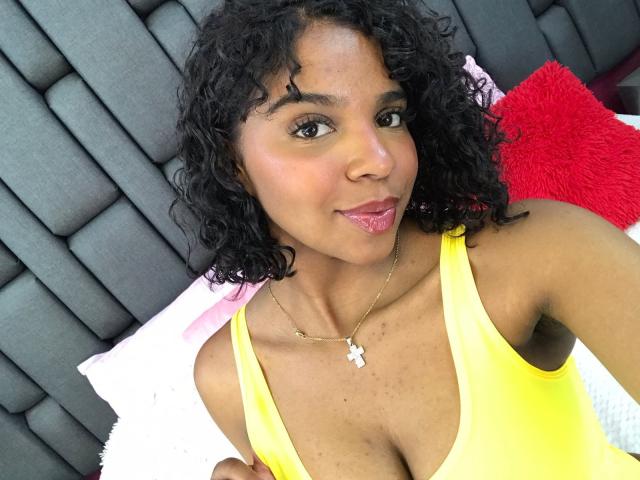 LisaMendez with curly black hair, brown skin, wearing a yellow tank top, showing her tits, pink lipstick, and a cross necklace, smiling at the camera.